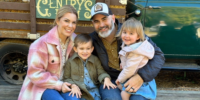 Tasha Layton wearing a pink plaid jacket, a printed blouse and jeans posing with her smiling husband and two children