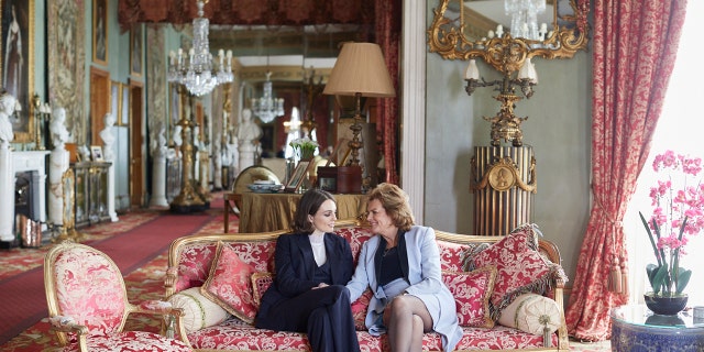 Lady Violet Manners in a navy blazer and pants with a white turtleneck sitting on a pink couch with her mother wearing a light blue blazer and a matching skirt.