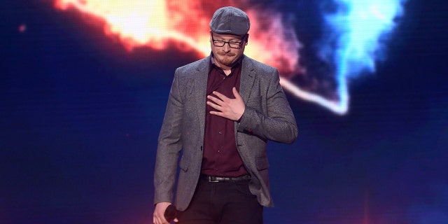 Art Garfunkel Jr. wearing a gray blazer and matching hat with a burgundy shirt performing on stage
