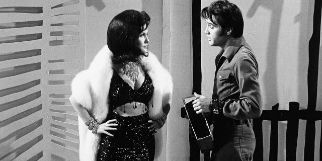 A model wearing a bejeweled bra top and skirt in a white fur coat talking to Elvis Presley in a denim suit