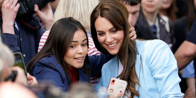 Kate Middleton in a light blue blazer and white blouse taking a selfie with a woman in a navy blazer