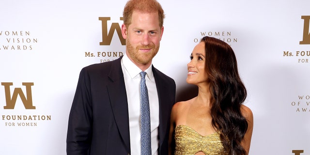 Prince Harry wearing a navy suit and a white shirt with a light blue tie posing next to Meghan Markle wearing a strapless gold dress