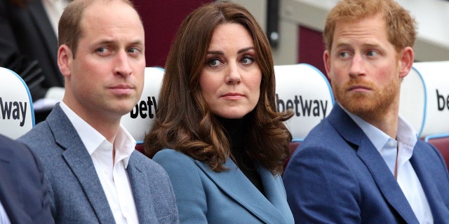 Kate Middleton wearing a blue blazer with a black sweater sitting in between Prince William and Prince Harry wearing various shades of blue and grey