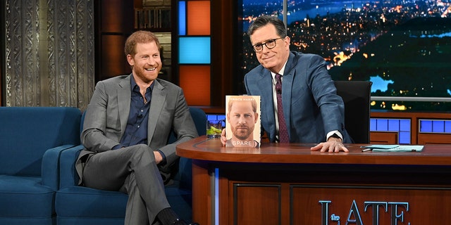 Prince Harry wearing a grey suit and blue shirt smiling sitting next to Stephen Colbert and his memoir Spare