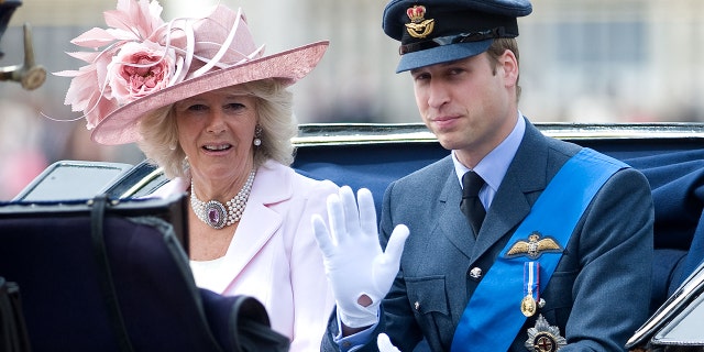 Prince William in a military suit sitting next to Queen Camilla in a pink dress and matching hat