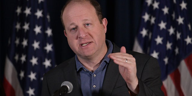 Jared Polis speaking with American flags in the background