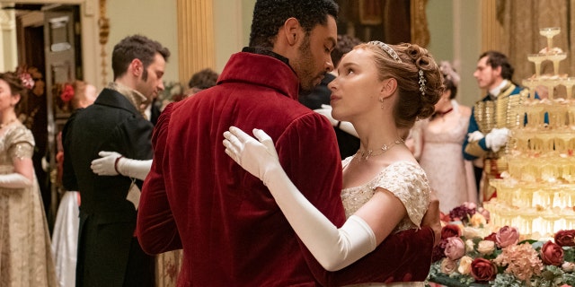 The Duke of Hastings wearing a burgundy coat dancing with Daphne Bridgerton wearing a lace white dress