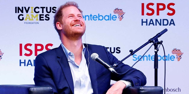 Prince Harry smiling wearing a navy blazer and a open button shirt