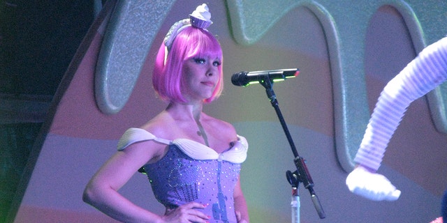 Tasha Layton looking serious wearing a hot pink wig and a lavendar and white dress on stage standing in front of a mic