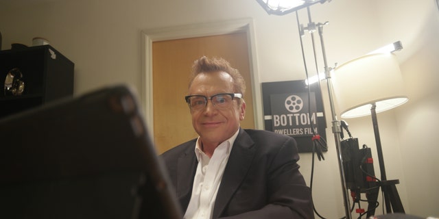 Tom Arnold sitting at a desk in character
