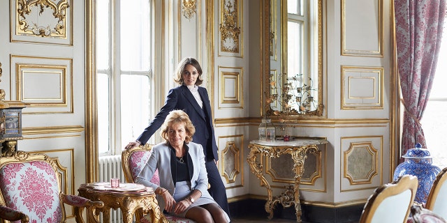 Lady Violet Manners wearing a navy blazer and pants with a white turtleneck posing with her mother, wearing a light blue blazer and skirt with a dark blouse