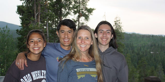 Wendi Lou Lee wearing a dark grey/blue sweater smiling next to her husband and two children in the woods