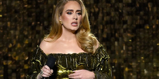 Adele in a green dress sits at the piano, holding a microphone and looking out at the crowd in London