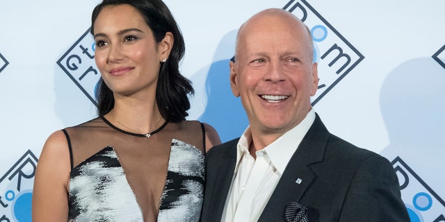 Emma Heming Willis looks to her right in a blue dress with black mesh as Bruce Willis looks to his left wearing a dark suit on the carpet