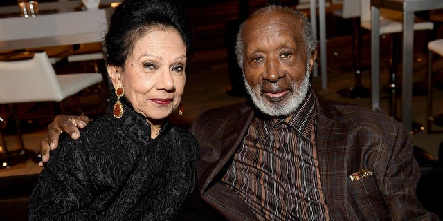 Jacqueline Avant soft smiles in a black outfit next to Clarence Avant in a brown jacket and printed shirt at a SAG-AFTRA Foundation Patron of the Artists Awards