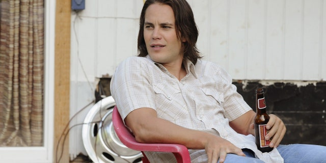 Taylor Kitsch as Tim Riggins in "Friday Night Lights" sits on a red chair in a white shirt and blue jeans