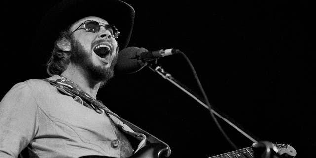Black and white photo of Hank Williams Jr. passionately singing into the microphone wearing a black hat and sunglasses on stage