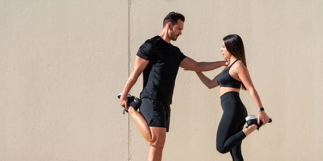 A tall man and short woman in gym attire stretch together.