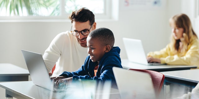 Male teacher helps a young boy with computer-based learning in a classroom setting.