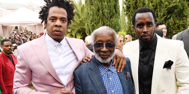 Jay-Z in a pink suit puts his arm around Clarence Avant and Diddy puts in a white suit and black shirt puts his arm around Clarence on the other side