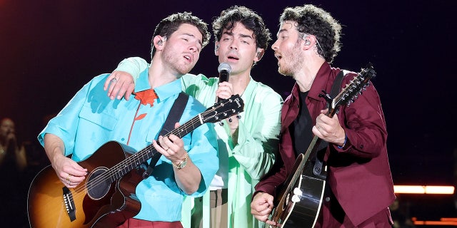 Nick Jonas playing the guitar leans back into Joe Jonas singing with Kevin Jonas, also playing the guitar leaning in to sing while on stage