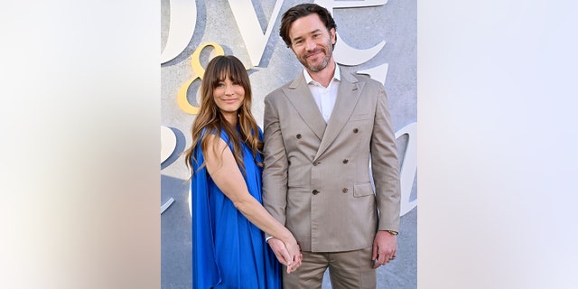 Kaley Cuoco in a long sleeveless blue silk dress holds hands with Tom Pelphrey on the carpet