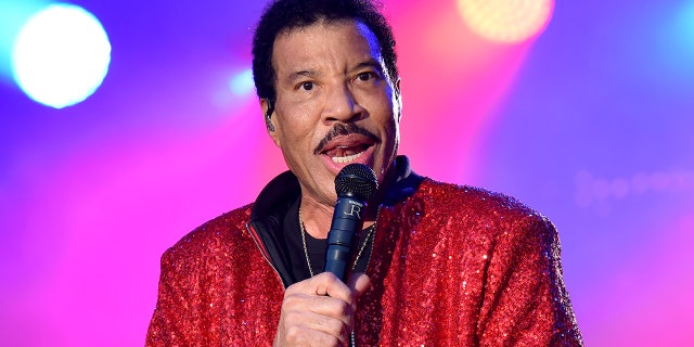 Lionel Richie speaks into the microphone on stage wearing a sparkly red jacket