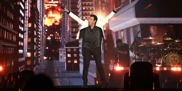 Lionel Richie wears a white jacket and black pants and shirt and puts his arms in the air on his tour with Earth, Wind & Fire