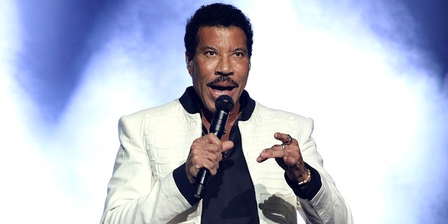 Lionel Richie performing on stage wearing a white jacket and black shirt in front of a blue and white background