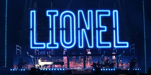 Lionel Richie performing on stage with his name outlined in blue lights on the screen behind him