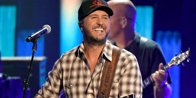 Luke Bryan in a checkered shirt and black cap plays the guitar at the ACM Awards