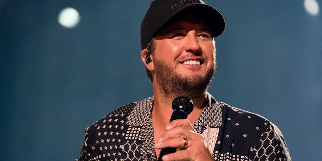 Luke Bryan in a patterned dark shirt and black cap looks up to his left while holding a microphone