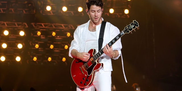 Nick Jonas in all white plays the guitar on stage and looks down at Yankee Stadium