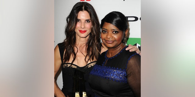 Sandra Bullock in a black dress puts her arm around Octavia Spencer in a dark dress with a blue stripe