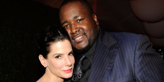 Sandra Bullock leans against Quinton Aaron in a blue pinstripe suit and black shirt and tie