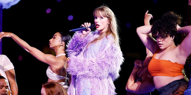 Taylor Swift hát trên sân khấu trong "The Eras Tour" trong chiếc váy lấp lánh màu tím và áo khoác lông màu hoa oải hương