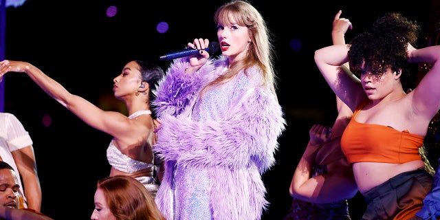 Taylor Swift sings on stage during "The Eras Tour" in a purple sparkle dress and feather lavender jacket