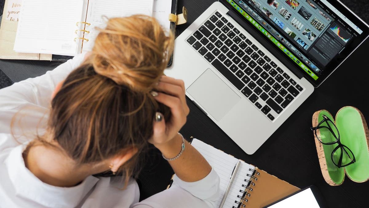 Woman sitting at her computer with her hands on her head and notebooks spread around.