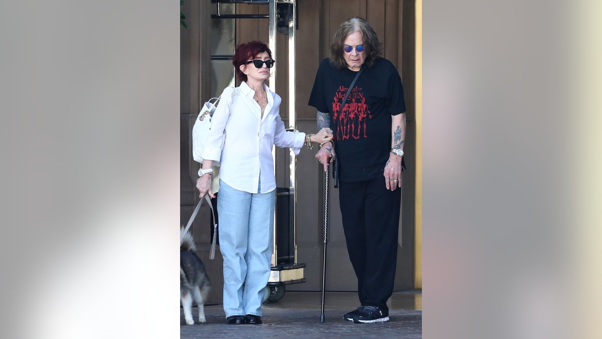 Sharon Osbourne wearing a white blouse and blue jeans walking alongside her husband Ozzy wearing black