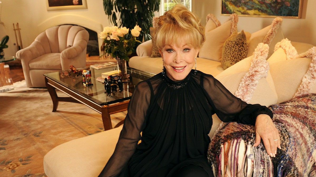 Barbara wearing a black dress with sheer sleeves sitting on a couch