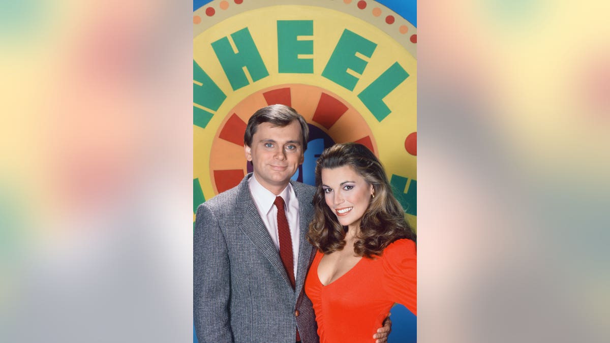 Pat Sajak and Vanna White stand in front of an old version of the Wheel of Fortune Wheel