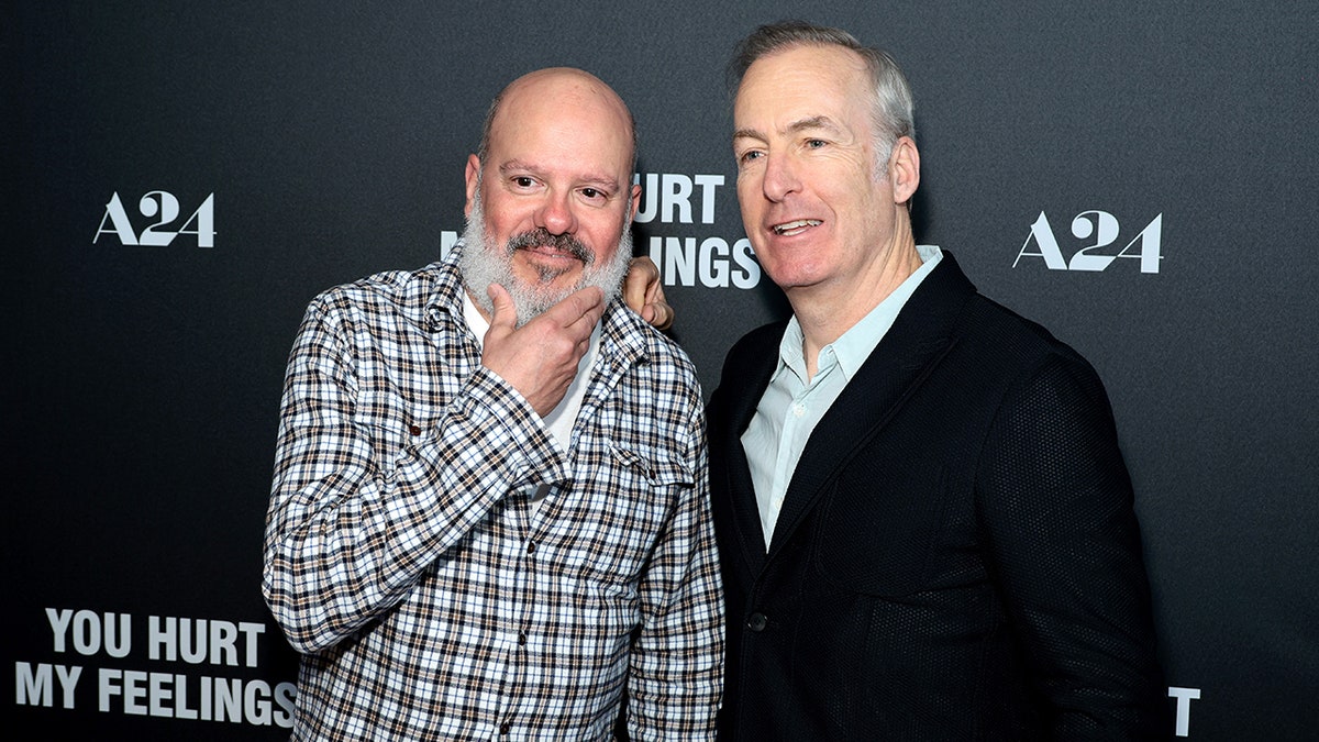 David Cross puts his hand under his chin indicating a sinister look while Bob Odenkirk in a black suit poses with him for a picture