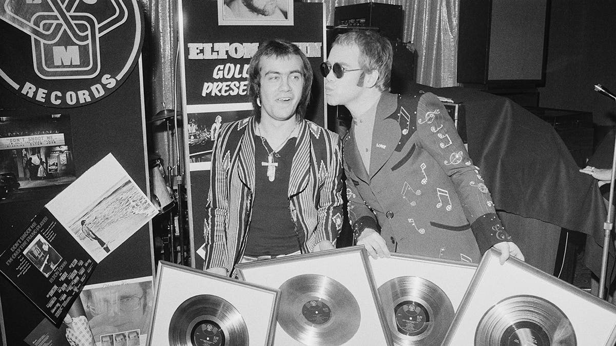 Elton John and Bernie Taupin holding a bunch of gold records