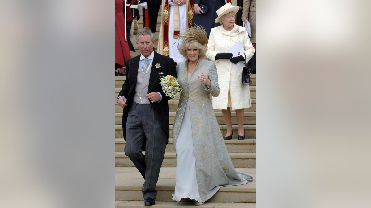 Camilla wearing a bridal gown next to Prince Charles in a suit with Queen Elizabeth behind in a creme dress