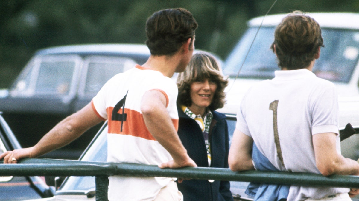 A young Camilla standing in front of two cars looking at Prince Charles and an unidentified man in polo uniforms