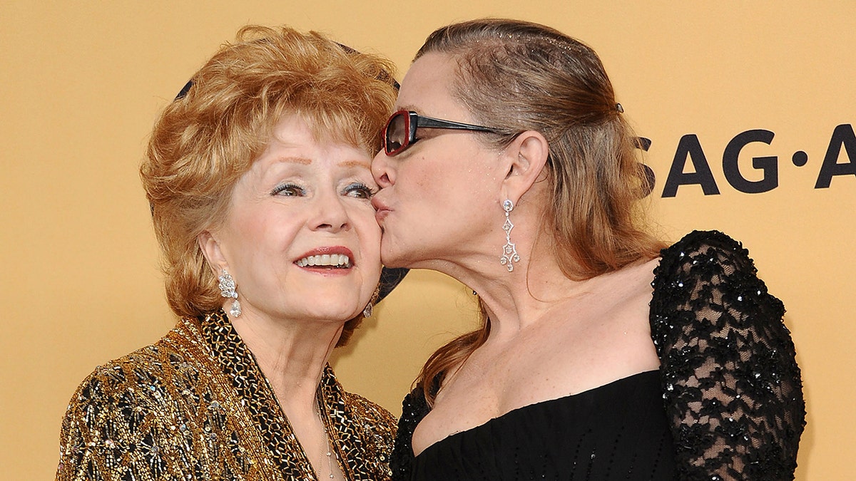 Debbie Reynolds in a gold dress getting kissed by her daughter Carrie Fisher in a black dress