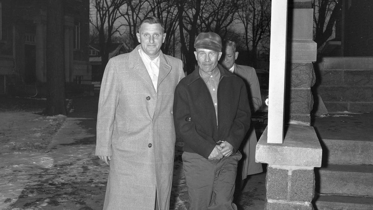 Ed Gein wearing a coat and a hat being escorted by police