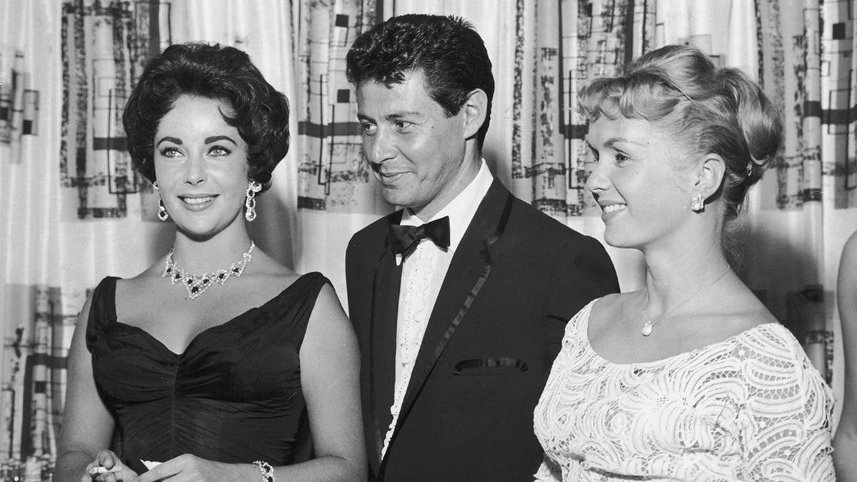 Foto em preto e branco de Elizabeth Taylor em um vestido preto e Eddie Fisher em um smoking e Debbie Reynolds sorrindo para uma foto