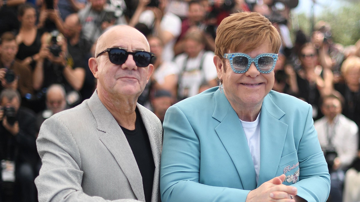 Bernie Taupin wearing a grey suit and a black shirt standing next to Elton John in a blue blazer and white shirt