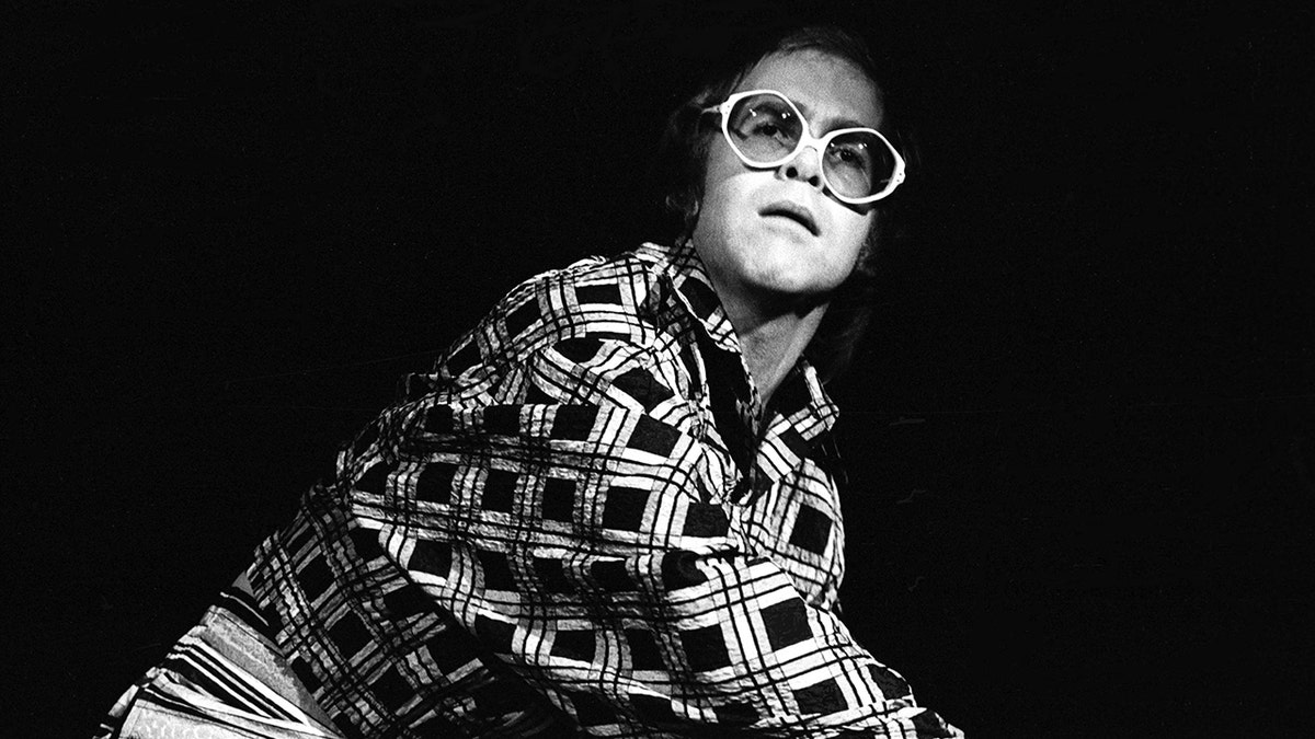 Elton John plays piano while wearing a multiprinted shirt